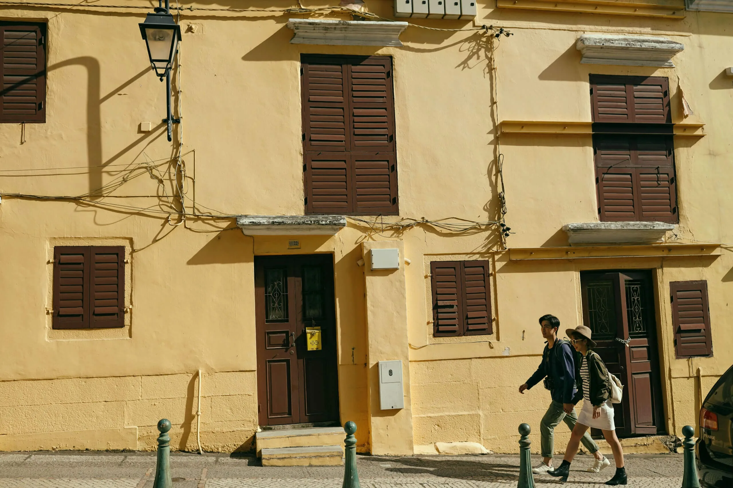Two travellers walking beside a sunlit yellow facade in Macau